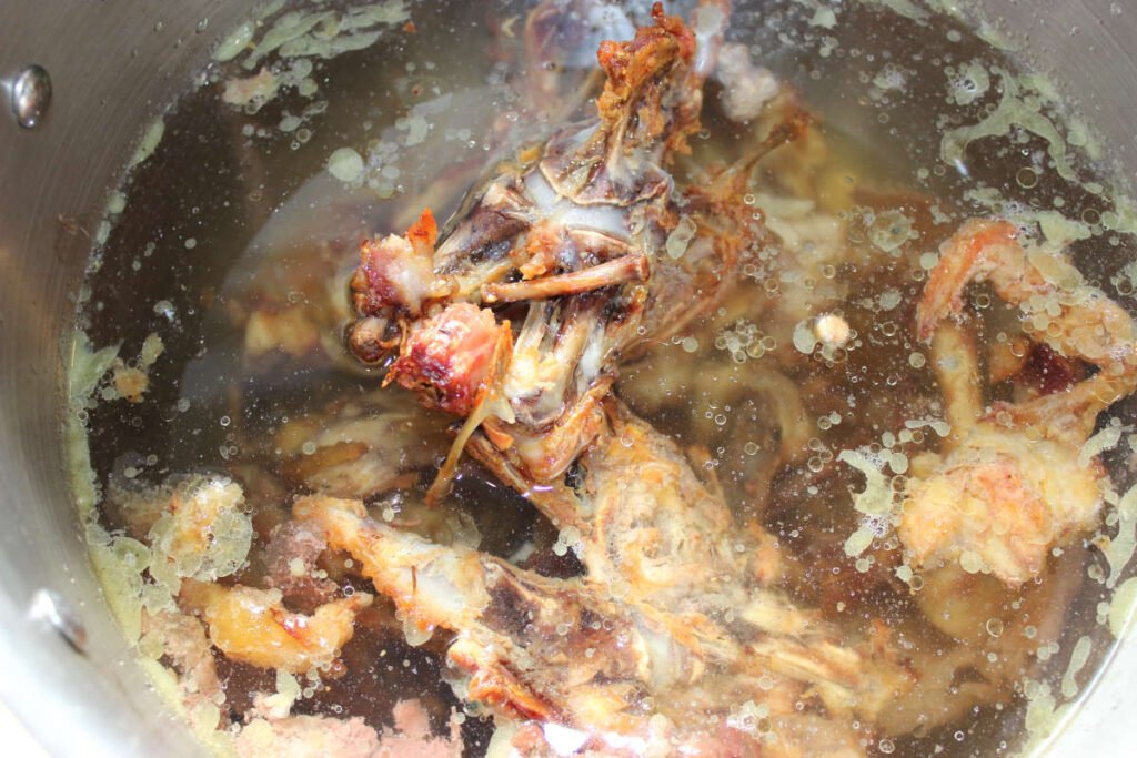 Close-up view of roasted chicken bones simmering in a large pot of water to make homemade chicken stock, with visible fat and impurities on the surface.