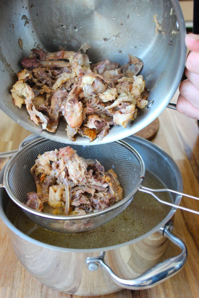 Cooked chicken bones draining in a fine-mesh strainer over a pot of freshly made homemade chicken stock.