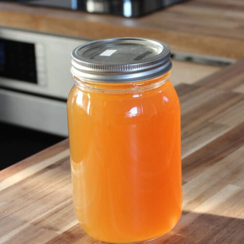 A glass jar filled with homemade chicken stock, sealed with a metal lid, sitting on a wooden kitchen countertop.