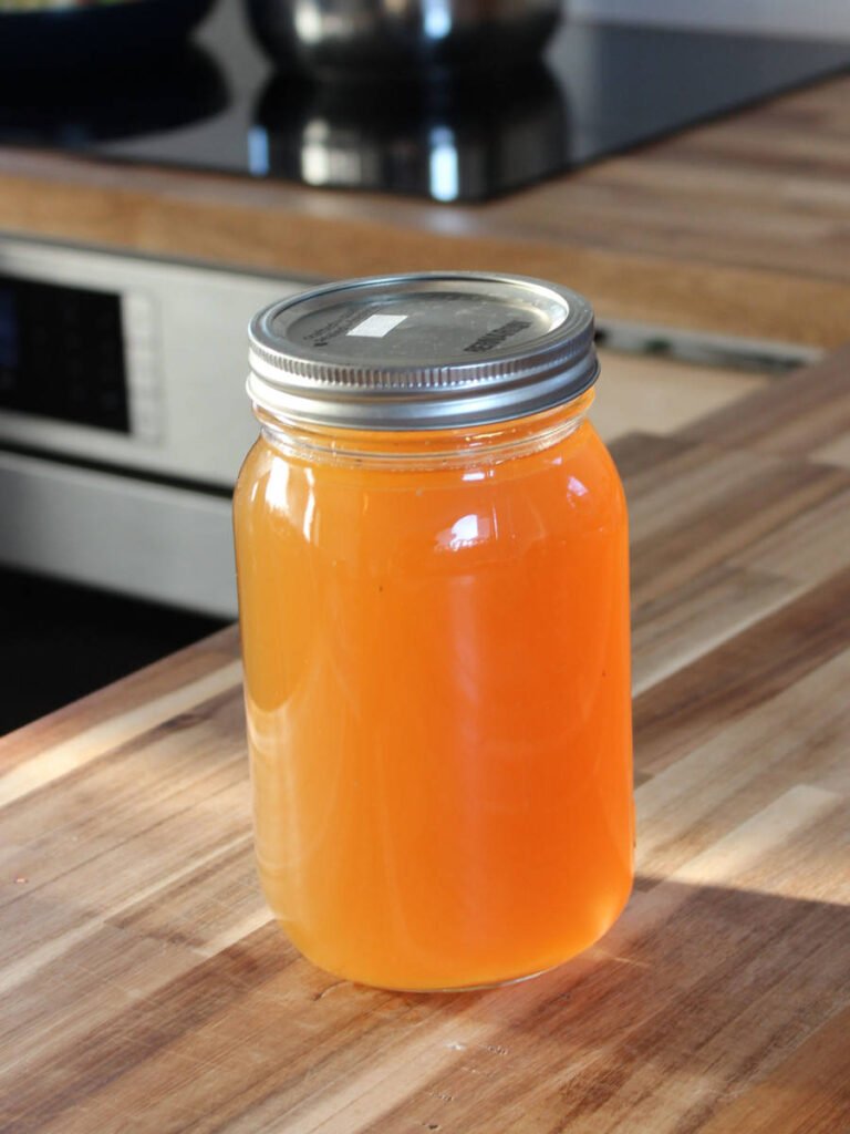 A glass jar filled with homemade chicken stock, sealed with a metal lid, sitting on a wooden kitchen countertop.