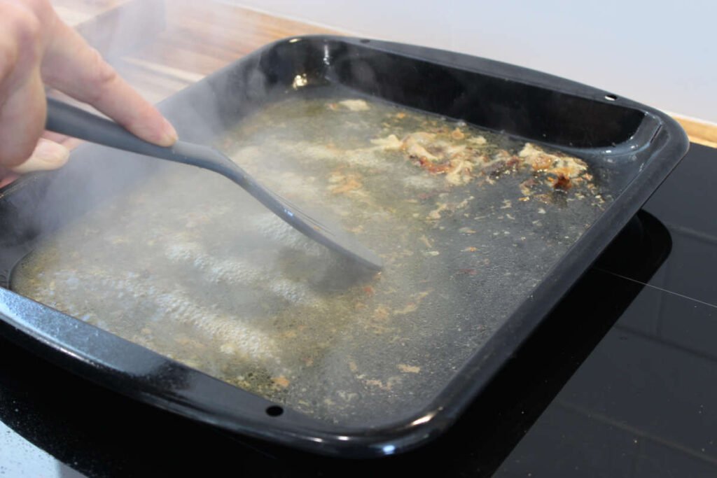 Hand using a spatula to scrape up flavorful browned bits from a roasting pan filled with hot water and chicken drippings to enhance homemade chicken stock.