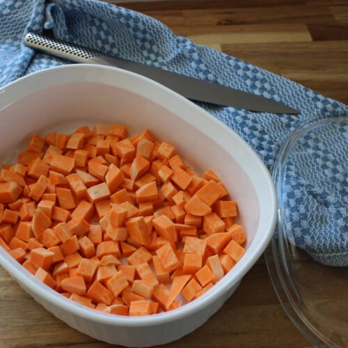 Bowl of cubed sweet potato ready to be microwaved sitting on a wooden counter next to a blue dish towel a knife and a lid