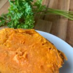 A whole sweet potato cooked in a microwave sitting on a white plate with parsley in the back. Microwaving sweet potato become super creamy and delicious.