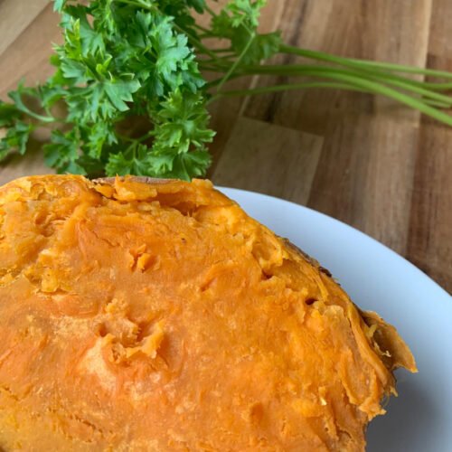 A whole sweet potato cooked in a microwave sitting on a white plate with parsley in the back. Microwaving sweet potato become super creamy and delicious.