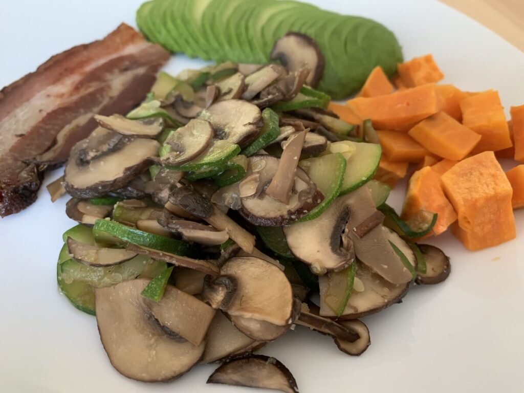 a white plate with an AIP breakfast, which consists of a slice of bacon, some fresh avocado, microwaved sweet potato and the star of the dish the sautéed mushrooms and zucchini
