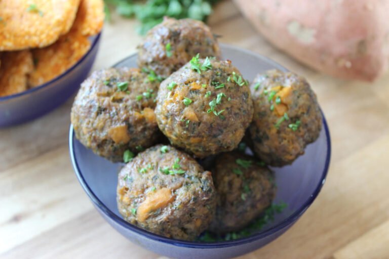 Meatballs in a blue bowl sitting on a wooden counter. In the background a sweet potato, parsley and a bowl of sweet potato patties