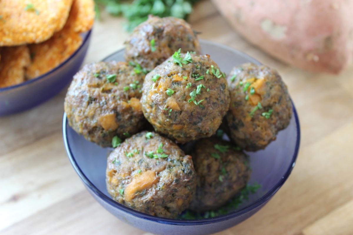 Meatballs in a blue bowl sitting on a wooden counter. In the background a sweet potato, parsley and a bowl of sweet potato patties