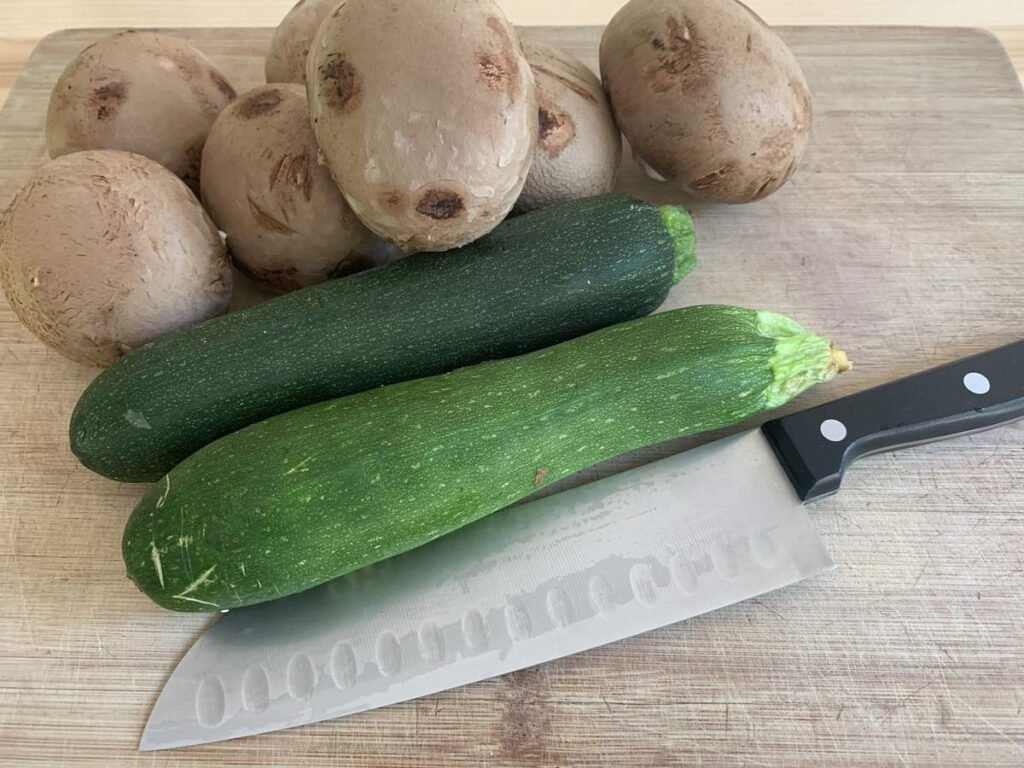 Some fresh mushrooms and zucchinis lying on a wooden cutting board with a kitchen knife next to them