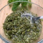 glass bowl of chimichurri and a teaspoon on a wooden counter with some fresh parsley in the back.