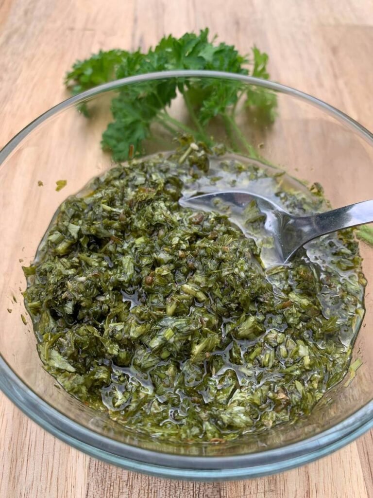 glass bowl of chimichurri and a teaspoon on a wooden counter with some fresh parsley in the back.