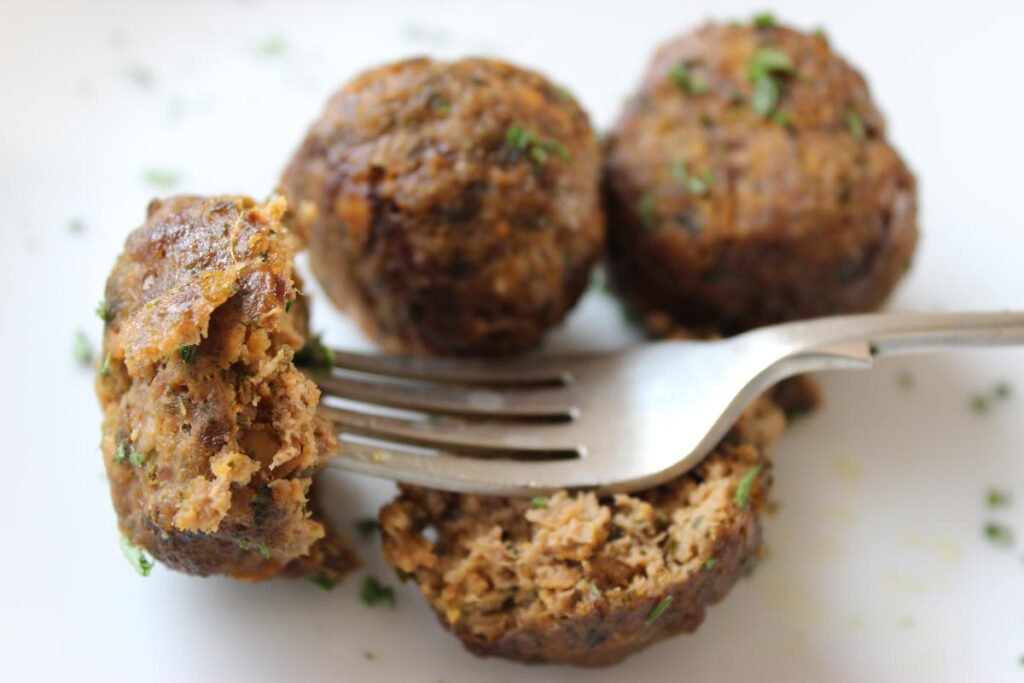 3 meatballs on a white plate. One meatball is broken apart and for is holding half of it.