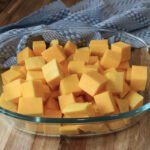 Glass dish with diced butternut squash on a wooden counter with a blue dishtowel in the background