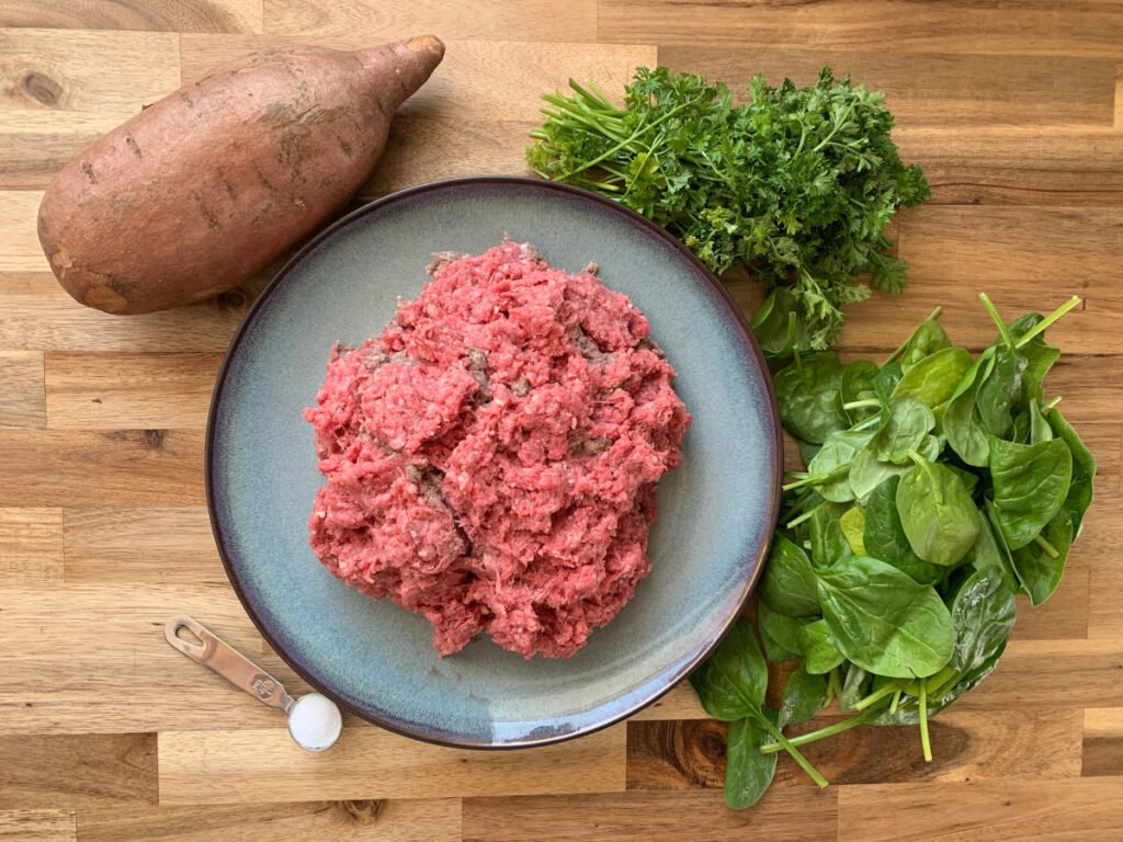 Ingredients for meatballs lying on a wooden counter. Ground beef on a blue plate, a sweet potato, salt in a teaspoon, spinach and fresh parsely