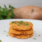 Gluten-free sweet potato cakes stacked on a plate, with fresh herbs and whole sweet potato for decoration
