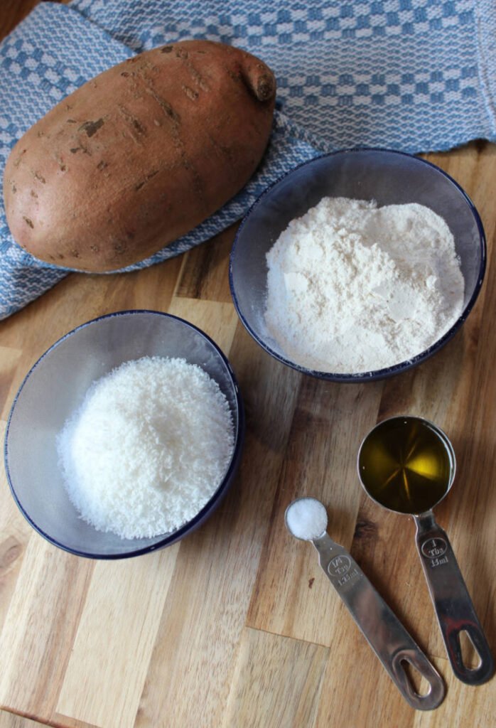 Sweet Potato Patty ingredients on a wooden cutting board. Ingredients include a sweet potato, olive oil, salt, cassava flour and optional desiccated coconut.