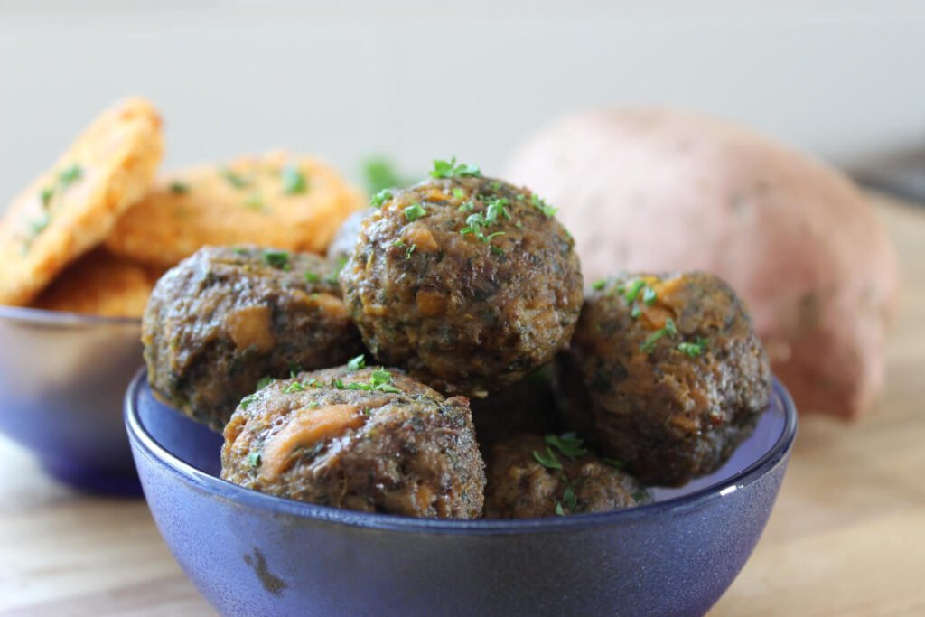 Sweet potato and spinach meatballs in a blue bowl sitting on a wooden counter. In the background a sweet potato, parsley and a bowl of sweet potato patties