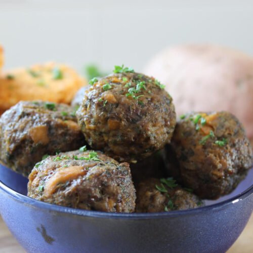 Sweet potato and spinach meatballs in a blue bowl sitting on a wooden counter. In the background a sweet potato, parsley and a bowl of sweet potato patties