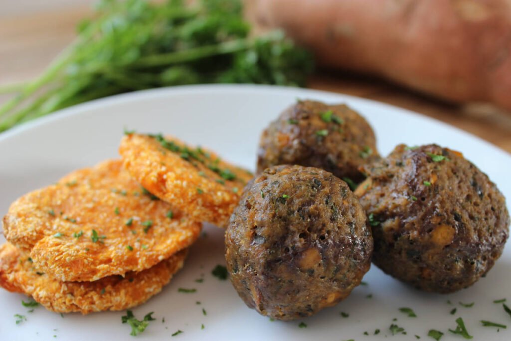 A white plate with three sweet potato and spinach meatballs and 3 sweet potato patties with a sprinkling of parsley. A sweet potato and fresh parsley in the background