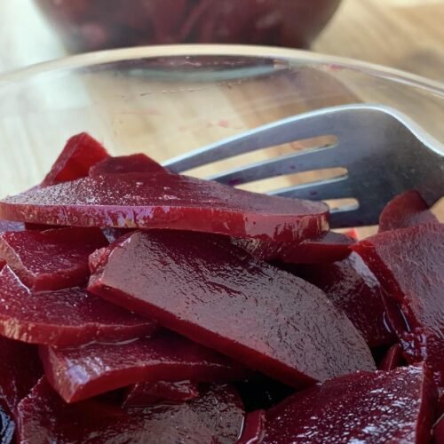 Glass bowl filled with AIP complient beetroot and a fork. In the background is a jar of pickled beets.