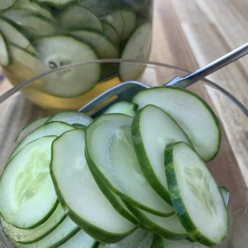 Glass bowl with fork, filled with sliced pickles. In the background is a glass jar filled with pickled cucumbers.
