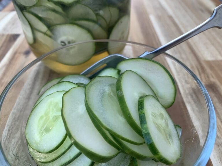 Glass bowl with fork, filled with sliced pickles. In the background is a glass jar filled with pickled cucumbers.