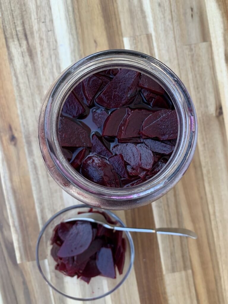 Glass jar filled with pickled beet. Next to it a small glass bowl with a fork and a portion of pickled beetroot for snacking on.