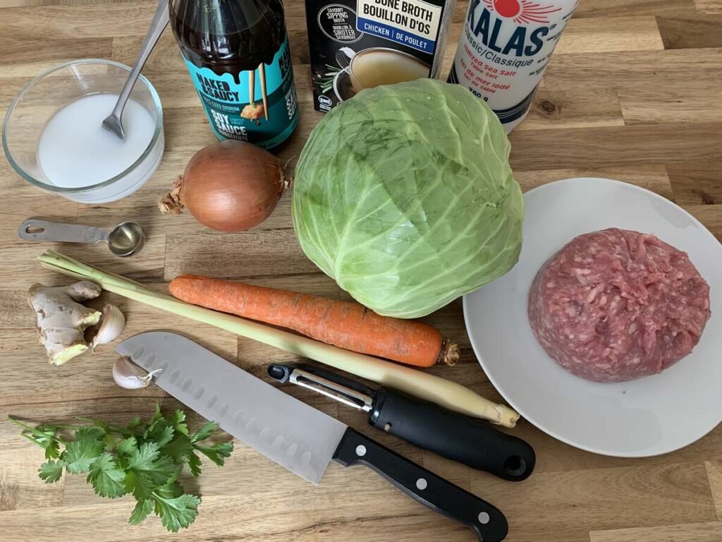 Ingredients for aisian-inspired ground pork AIP breakfast on a wooden cutting board. Main ingredients include ground pork, onion, garlic, lemongrass, ginger, cabbage and carrot.