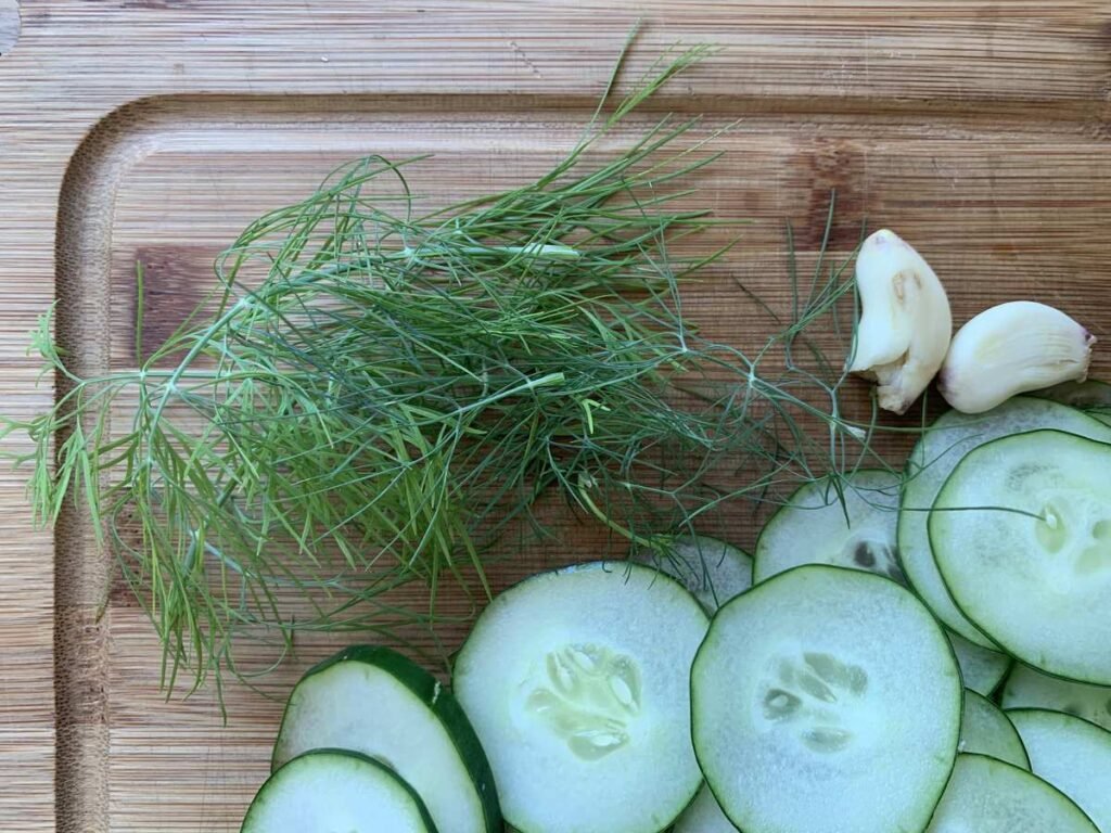 Cutting board with the freshingredients for AIP compliant pickled cucumbers: sliced cucumbers, 2 cloves of garlic and fresh dill