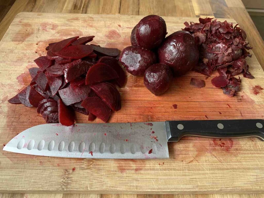 Cutting board with a knife. Behind the knife is a pile of sliced beetroot, some whole peeled beetroot and beetroot peel that has already been removed.