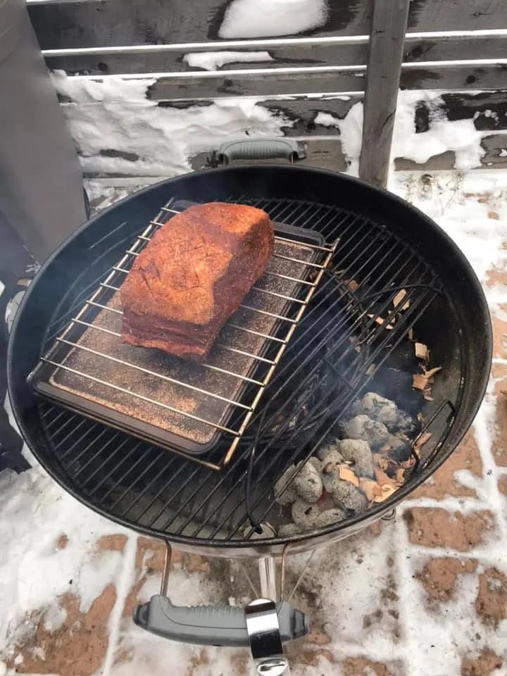 A kettle BBQ with a large piece of meat over a baking sheet on a grid over a BBQ snake. Woodchips can be seen at the start of the snake on the lit coals and some of the unlit coals.