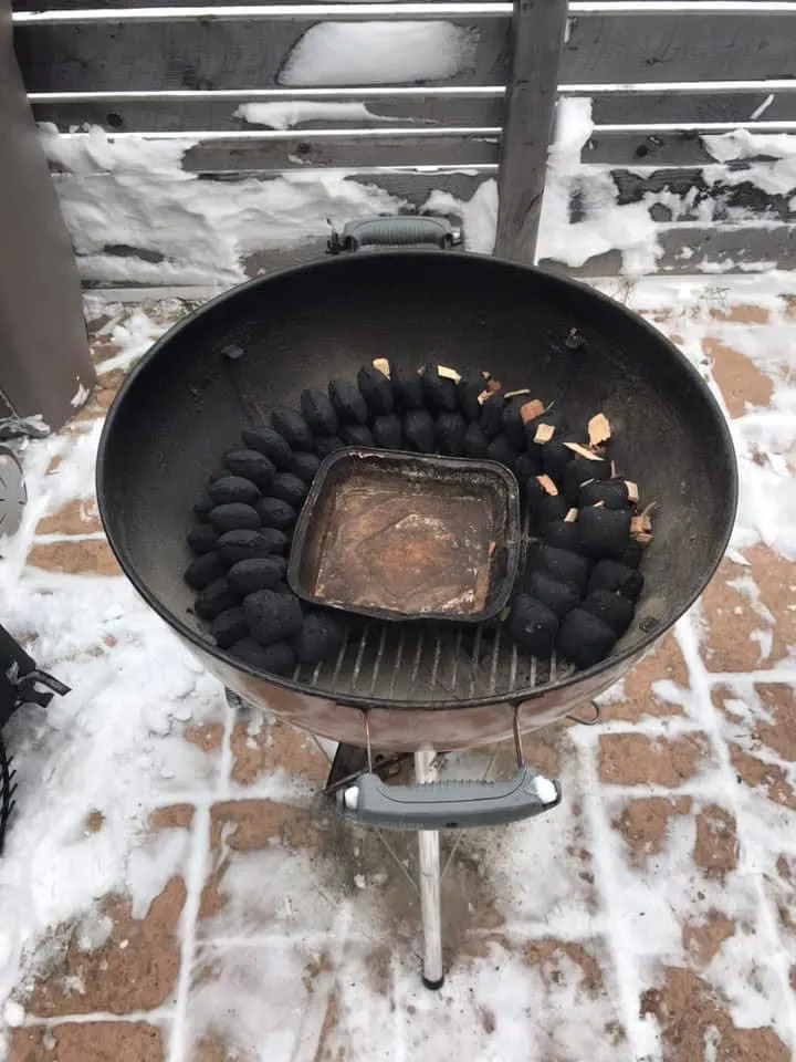 The BBQ snake is ready to be lit. Charcoal snake runs around the perimeter of the kettle with two rows at the bottom and one row on top. Wood chips are added to the start of the snake. There is a gap between the start and end of the snake to make space for adding lit coals to start the snake.
