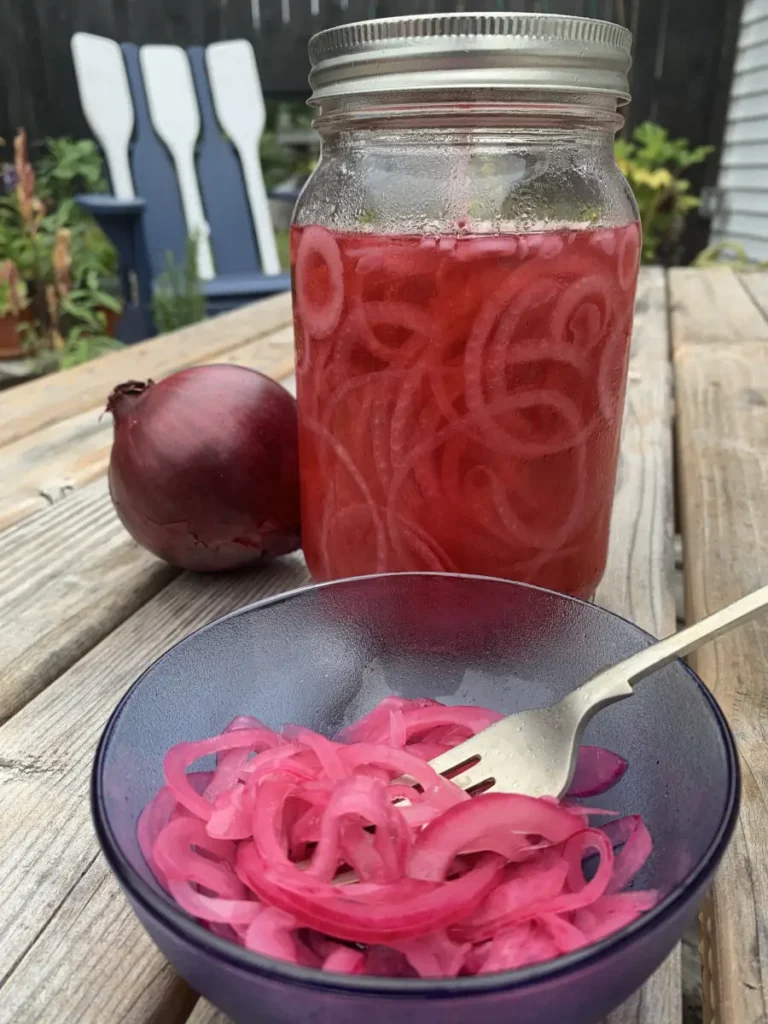 A bowl of pickled red onions standing in front of a jar of onions, a fresh red onion. Standing on a patio table with a blue chair and plants in the background.