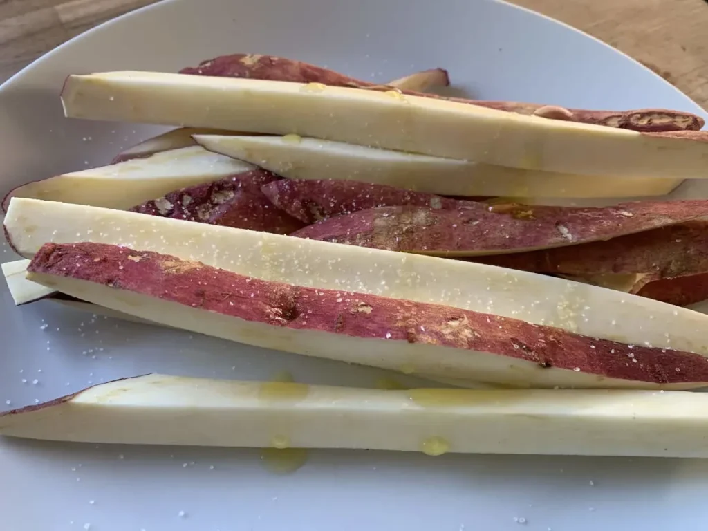 Sweet potato chips lying on a dinner plate with salt and olive oil drizzled over them, before going in the oven.