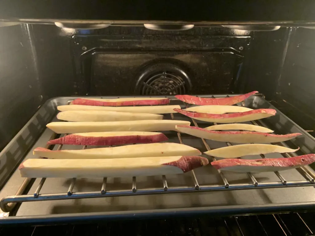 Sweet potato chips lying on a grid sitting over an oven tray in the oven, with the oven light on.