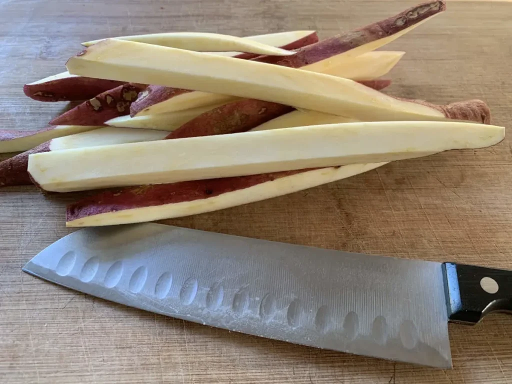 A santoku kitchen knife and a sliced up sweet potato lying on a wooden cutting board.