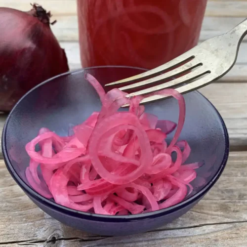 Bright pink pickled red onions in a blue glass bowl standing in front of a jar of pickled red onions and a fresh uncut red onion.