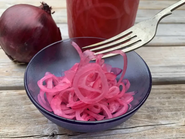 Bright pink pickled red onions in a blue glass bowl standing in front of a jar of pickled red onions and a fresh uncut red onion.