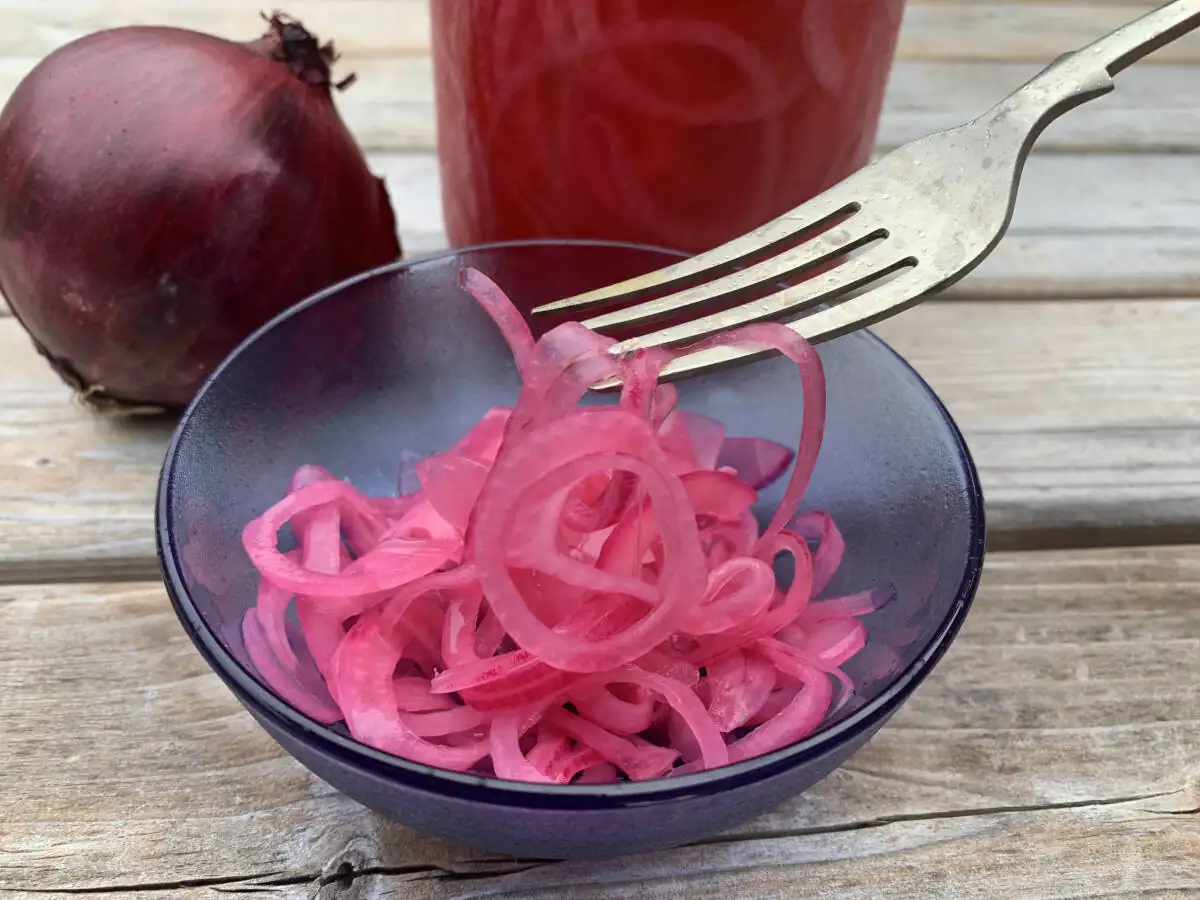 Bright pink pickled red onions in a blue glass bowl standing in front of a jar of pickled red onions and a fresh uncut red onion.