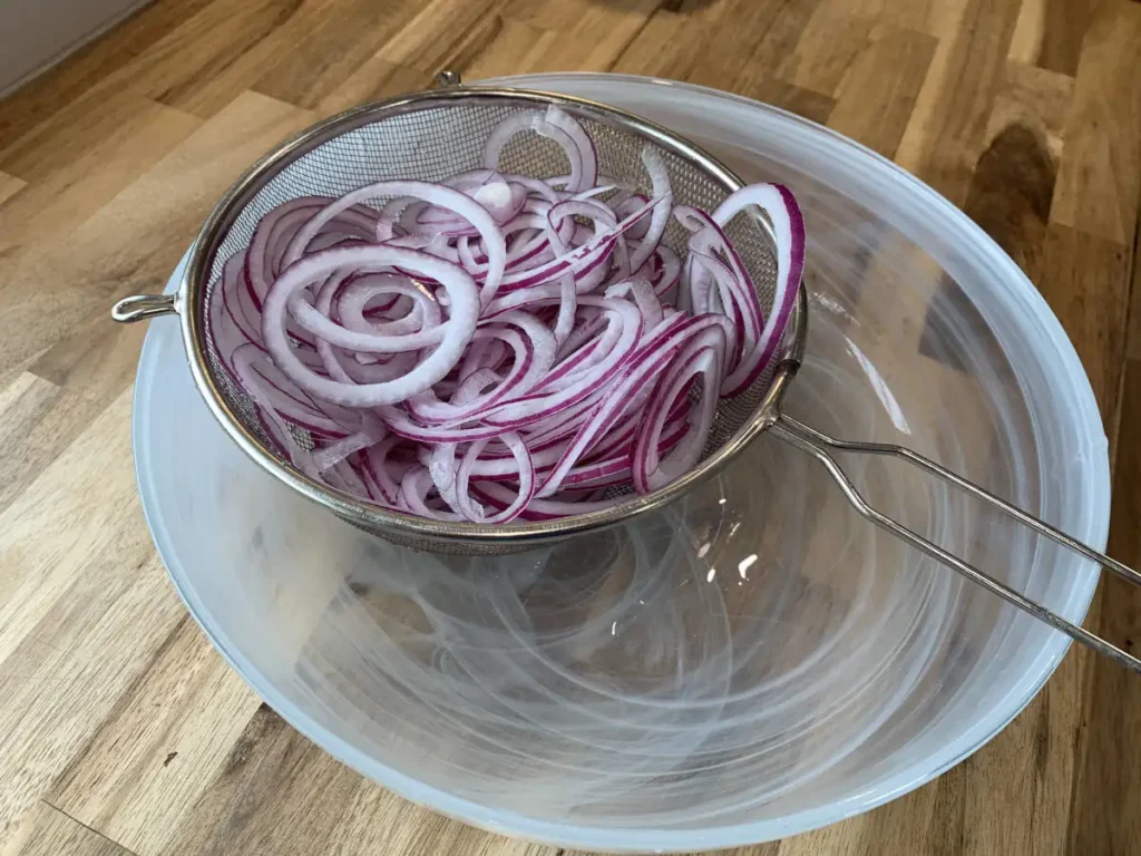 A sieve holding sliced red onions over a glass bowl, allowing the onions to drain.