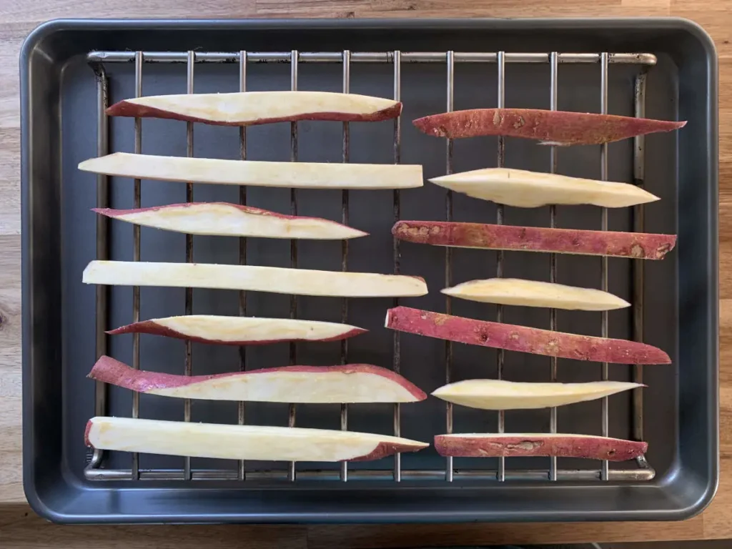 Sweet potato fries lying on a grid over an oven pan ready to be baked.