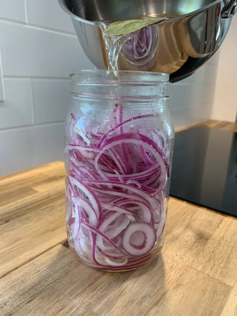A glass jar containing sliced red onions. From above a saucepan is pouring some pickling juice over the onions.