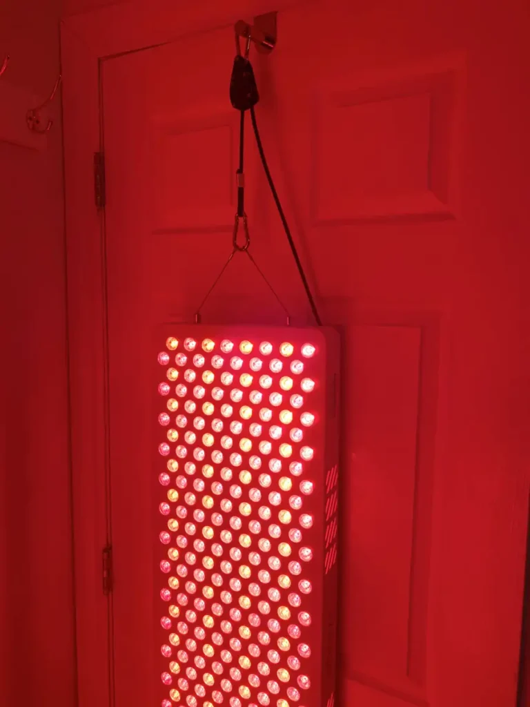 Red light therapy panel hanging on a door with pulley system for height adjustment. Red lights emmitting warm light in infrared and near infrared wavelengths.