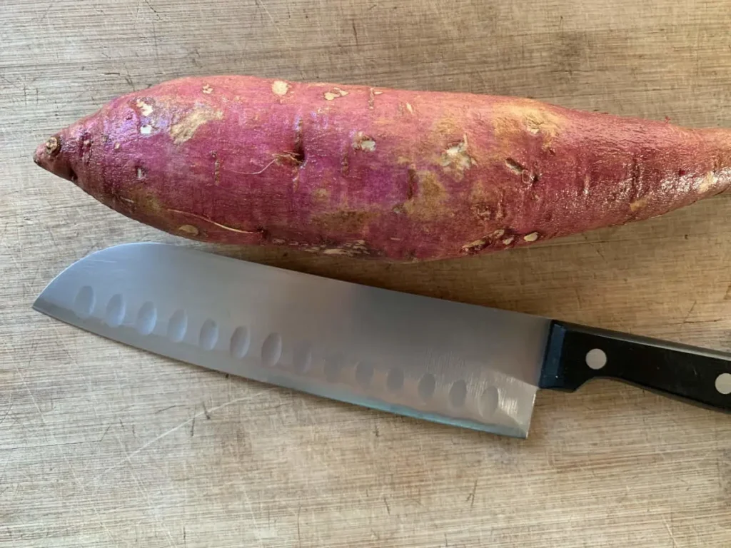 A kitchen knife and purple sweet potato lying on a wooden cutting board.