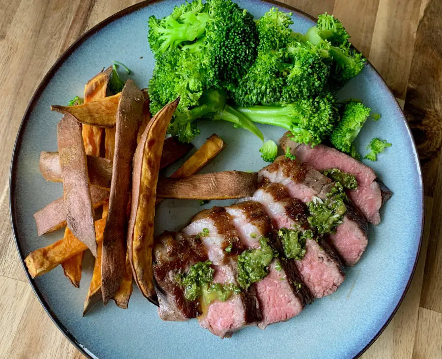 A plate sitting on a wooden counter with broccoli, sweet potato fries and a sliced up steak with chimichurri over it.