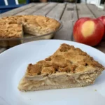 Slice of apple pie on a white plate sitting on a wooden table next toan apple pie in a glass pie dish and some fresh apples.