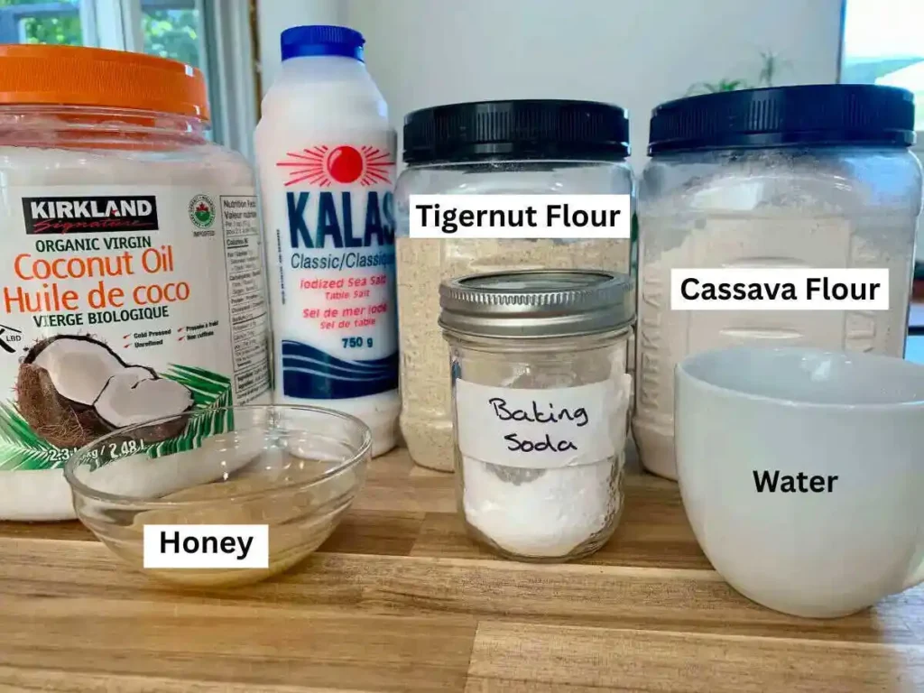 Ingredients for AIP apple pie crust lined up on a wooden counter. Coconut oil, salt, Tigernut flour, Cassava flour, Honey, Baking Soda and water.