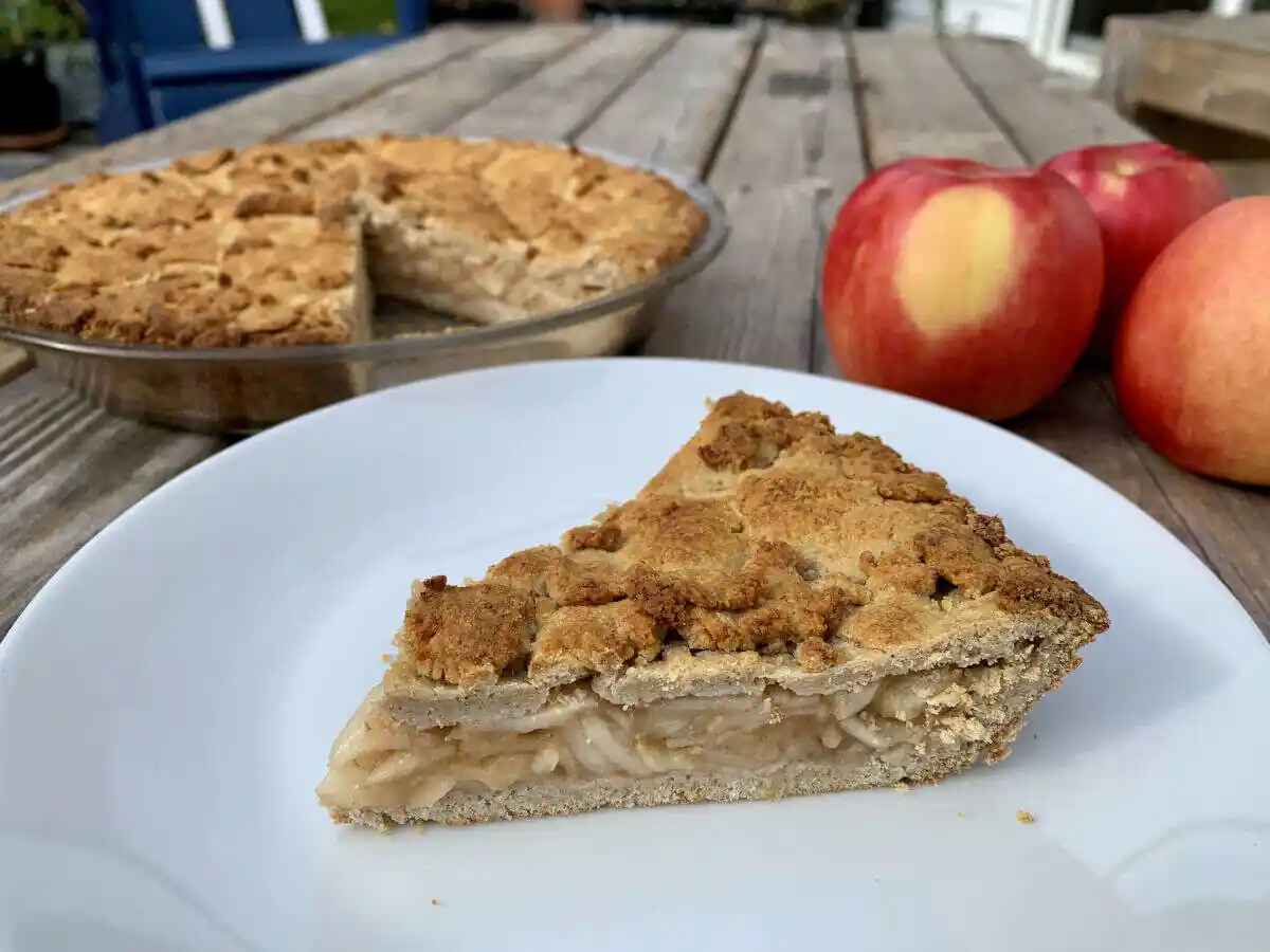 Slice of apple pie on a white plate sitting on a wooden table next toan apple pie in a glass pie dish and some fresh apples.