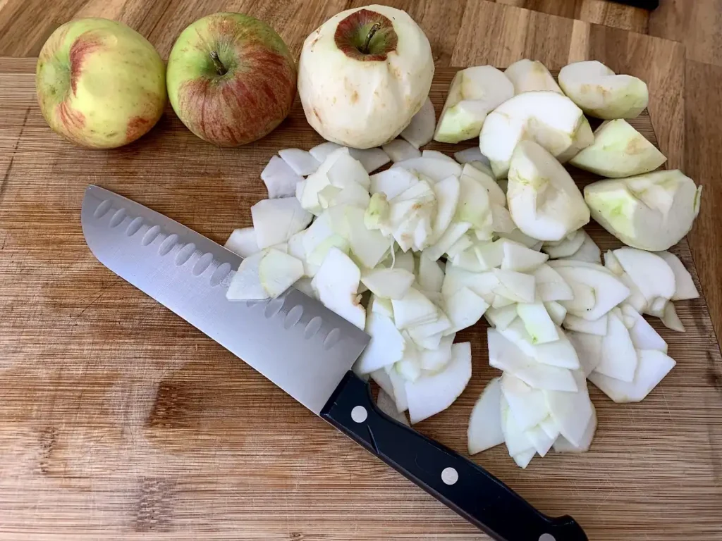 On a wooden cutting board, there are 2 whole apples one peeled apple and a few quartered and slices apples lying next to a kitchen knife.