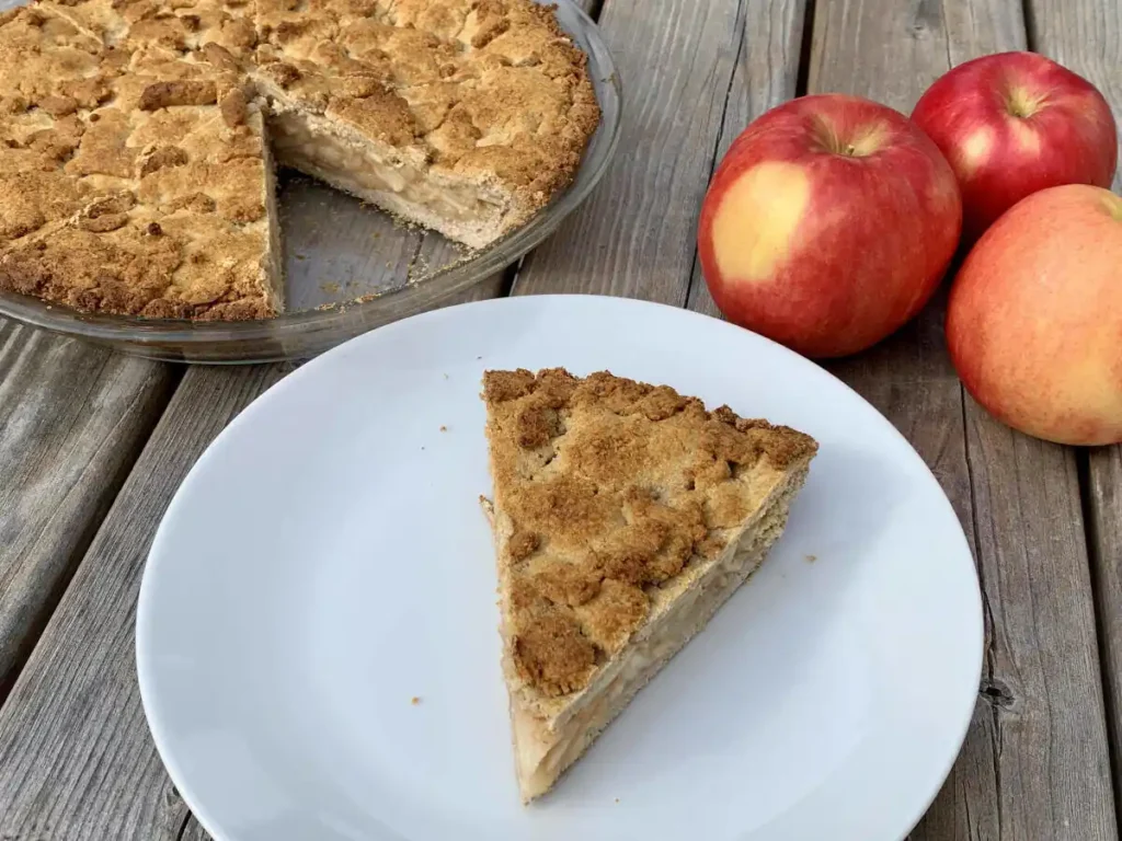 White plate with a slice of AIP apple pie sitting on a wooden table. Next to it is the rest of the pie in a glass dish and three fresh apples.