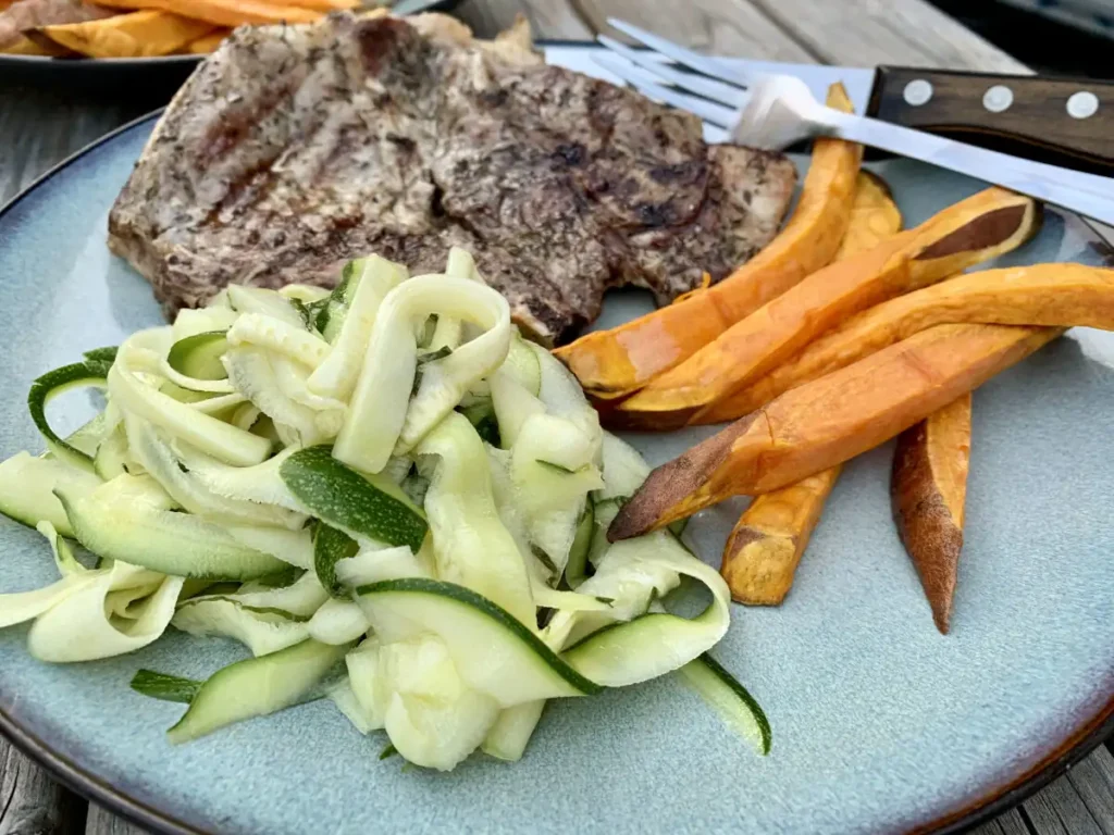 Uncooked zucchini salad served on a plate with grilled pork chops and roasted sweet potatoes fries outdoors